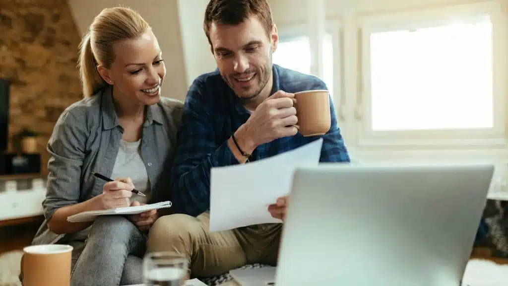 A man and woman look at papers together while sitting in front of a laptop.