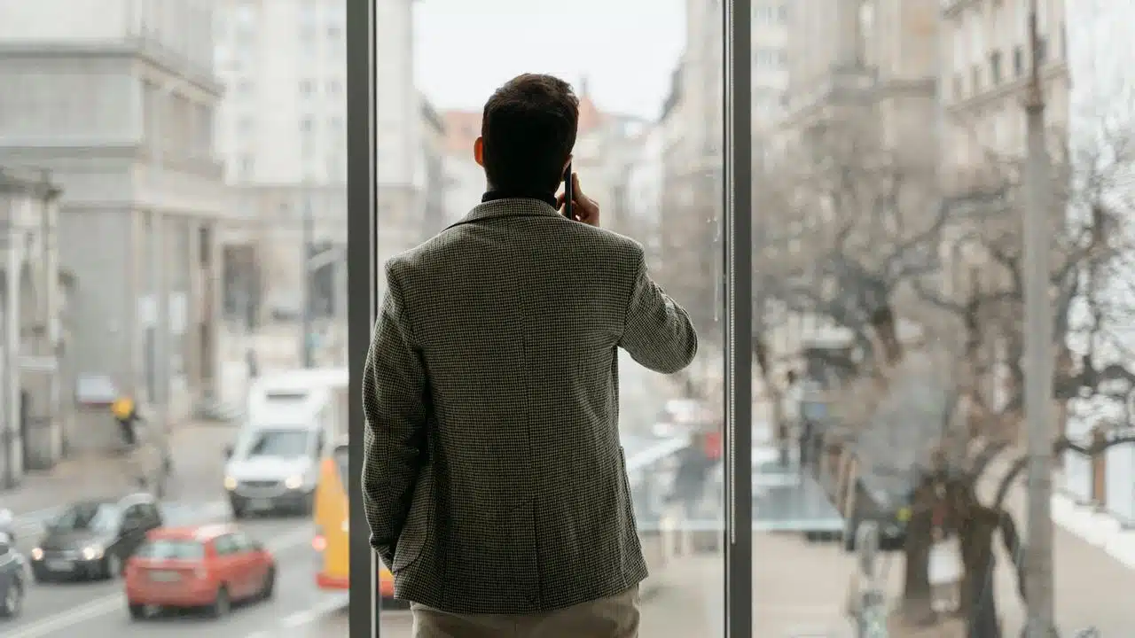 A man standing by a large window talking on his phone while looking outside.