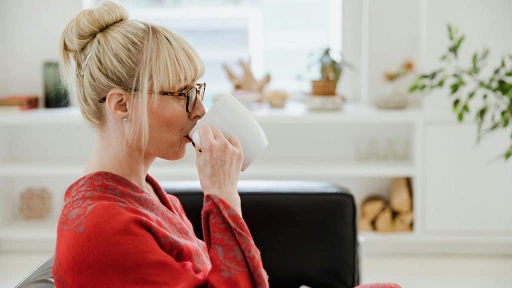 A woman wearing glasses and a red sweater sipping coffee from a white mug indoors.