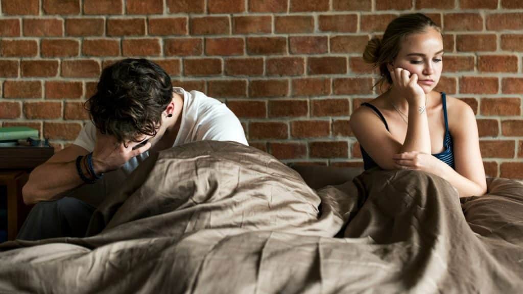 A man sits on a bed holding his head while a woman sits nearby looking away.