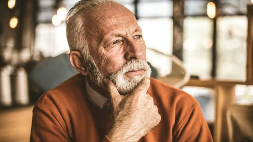 An older man with a white beard rests his chin on his hand while looking off-camera.