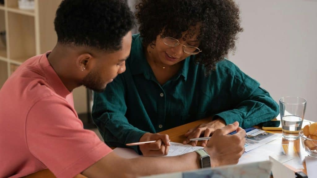 A man and woman sit at a table looking down at several papers and documents