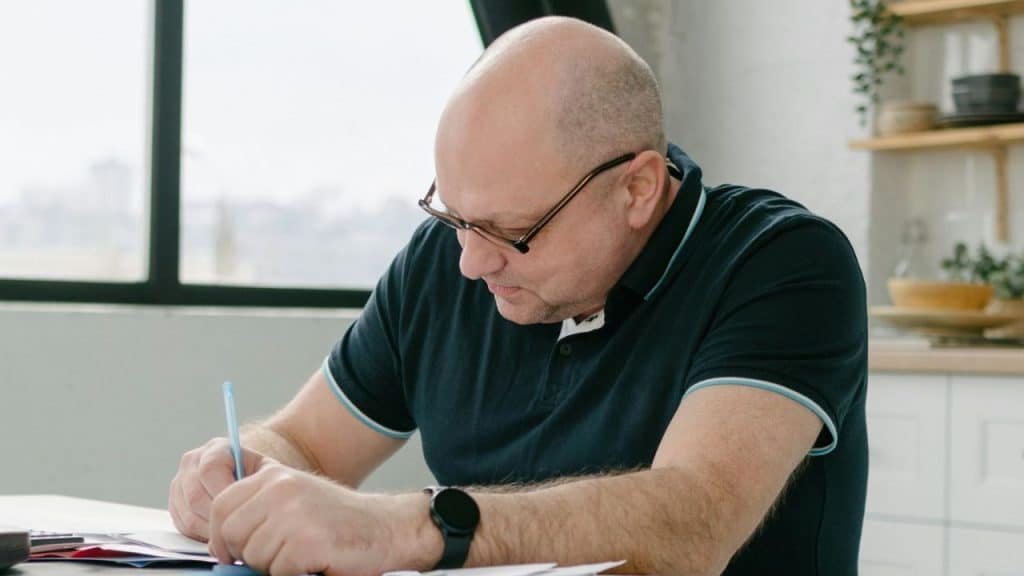 A bald man with glasses wearing a dark polo shirt writes on papers at a table.
