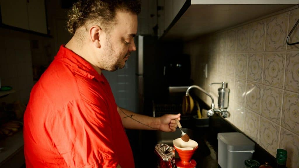 A man in a red shirt stands in a kitchen and spoons grounds into a filter.