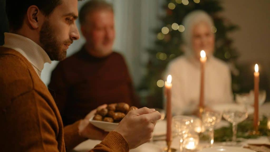 A man holding a bowl of food at a candlelit family dinner with two others in the background.
