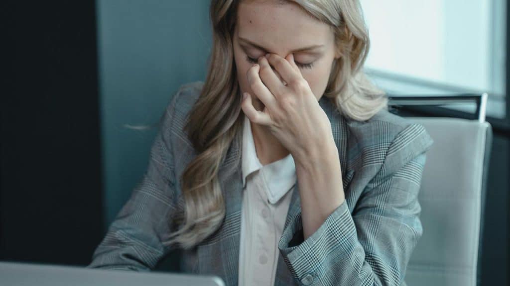 A woman sitting at a desk rubbing her eyes while looking tired in front of a laptop.