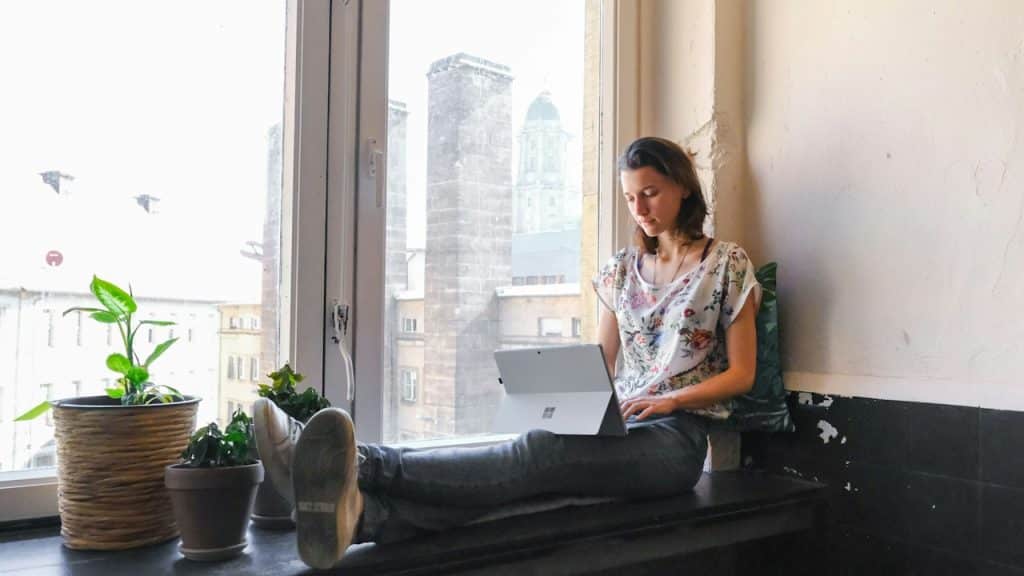 A woman using her laptop while sitting on a bench by a window.
