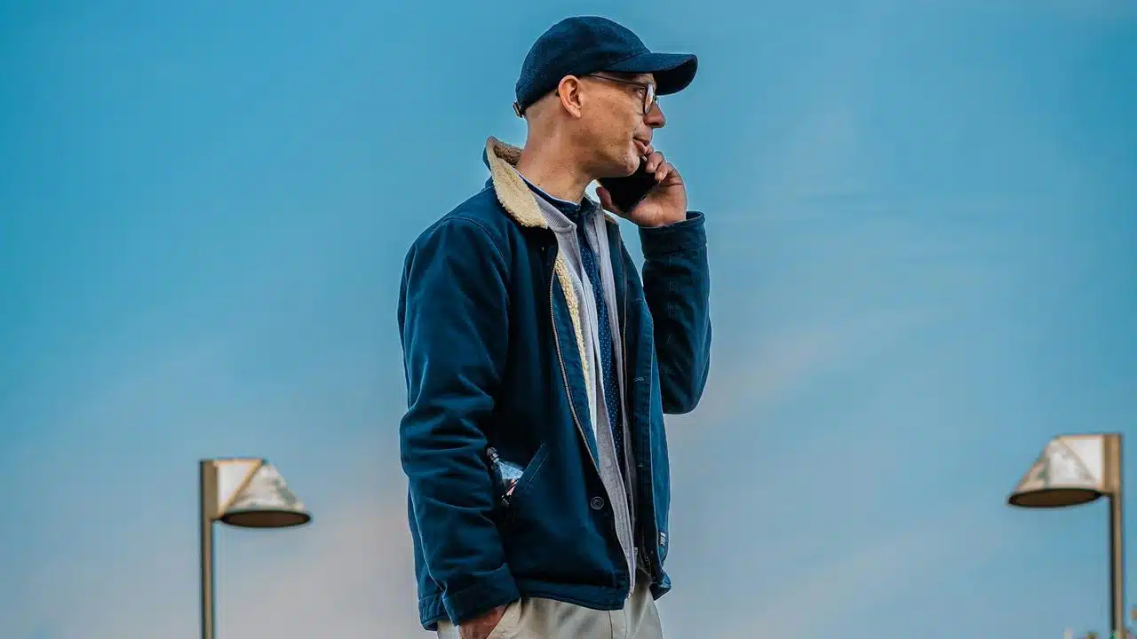 A man wearing a cap and glasses talking on his phone against a blue sky.