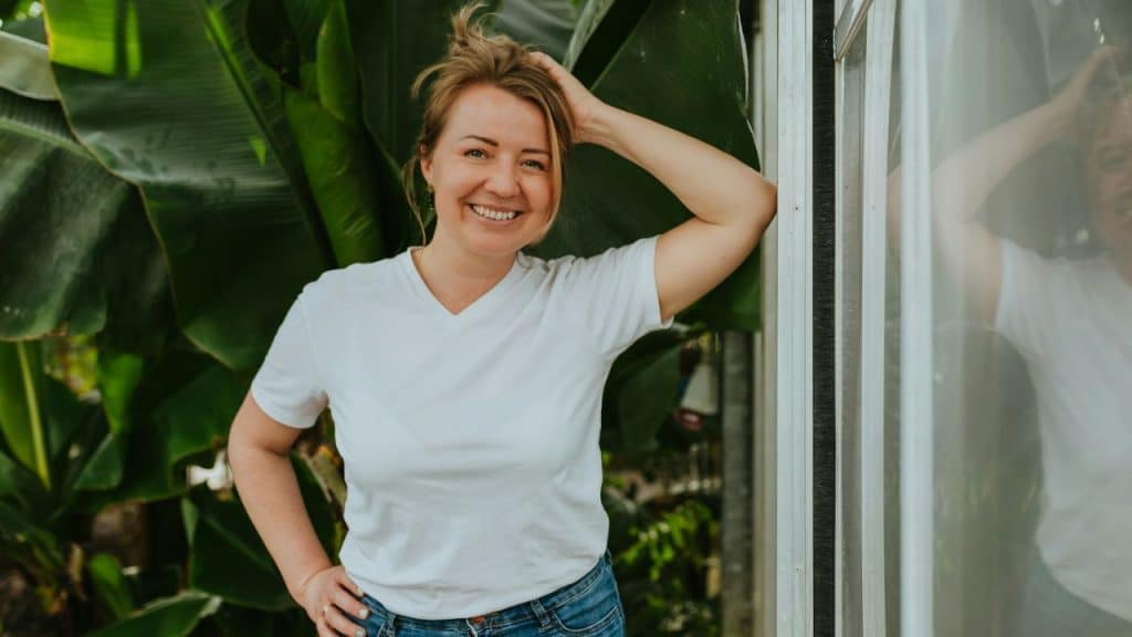 A woman in a white t-shirt and jeans smiles while leaning against a glass wall.