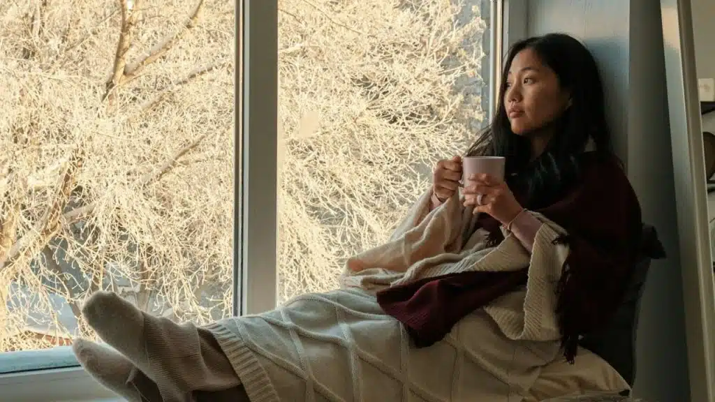 A woman wrapped in a blanket sits by a window holding a mug and looking out.
