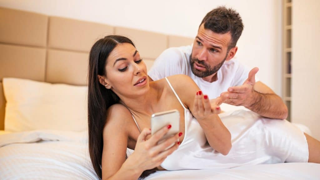 A man asking a woman about her phone while she gestures with her hand annoyingly as they lie on a bed.