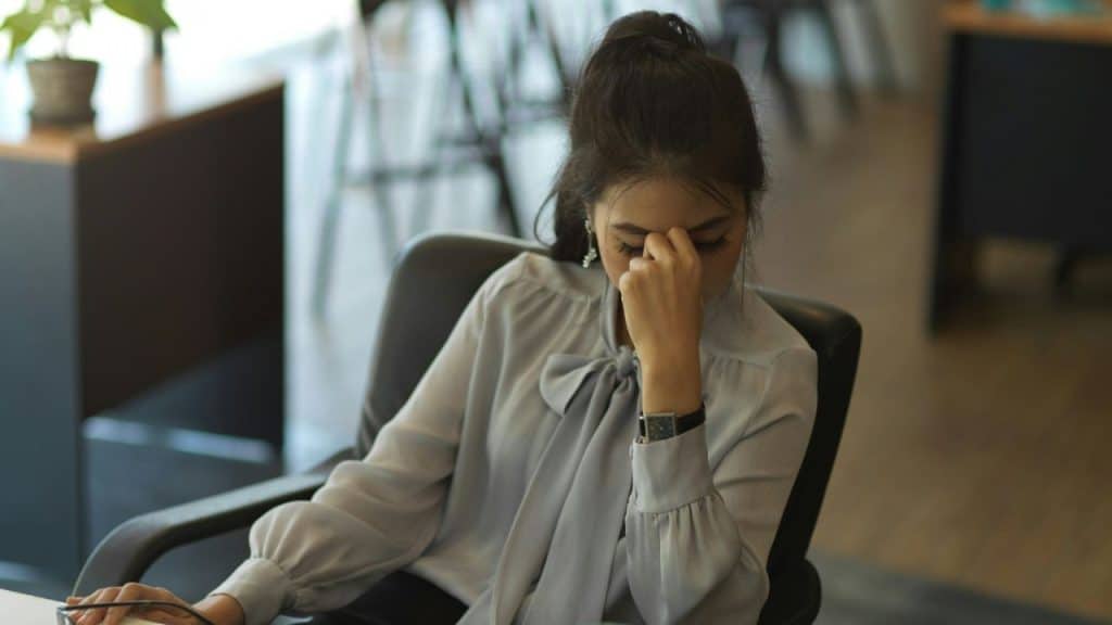 A woman in a grey blouse with a bow sits in a chair, pinching the bridge of her nose.