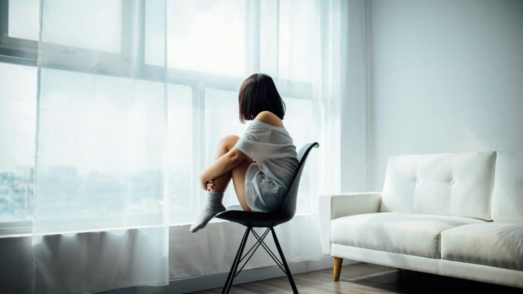 A woman is sitting alone in a modern, brightly-lit room, facing away toward a window with sheer curtains, with her knees hugged to her chest.