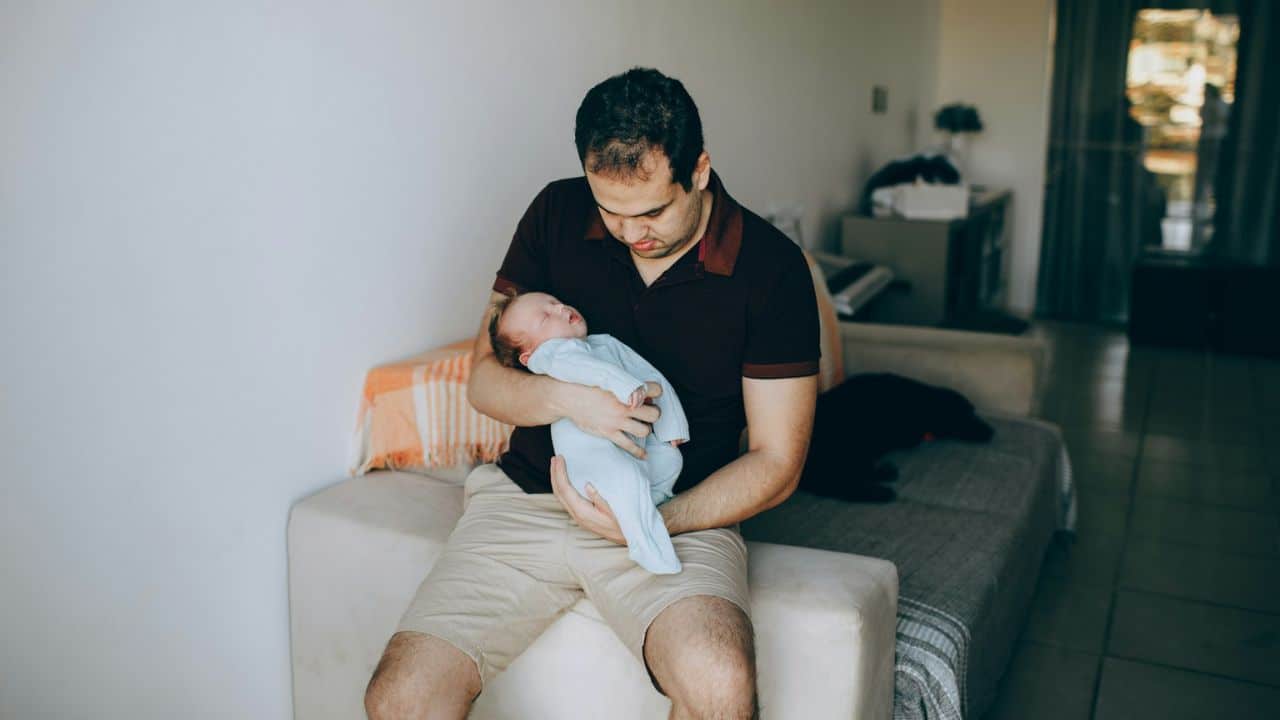 Man in a polo shirt and shorts sitting down while holding a sleeping baby.