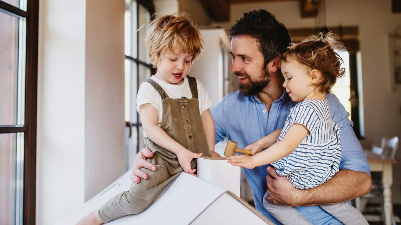 Father sitting with two small children building a cardboard structure indoors.