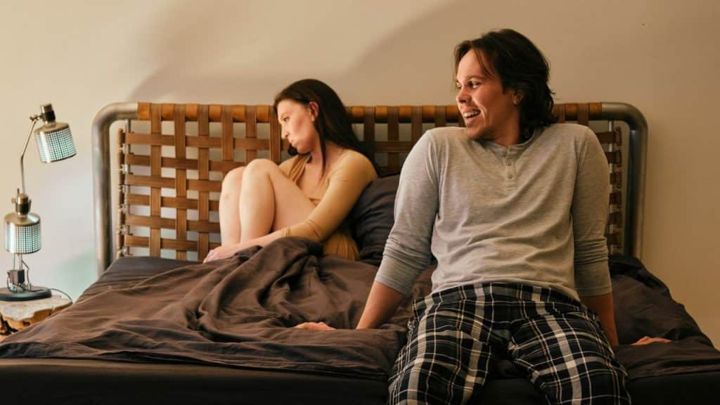 A man is smiling at a woman who is sitting away from him on a bed.