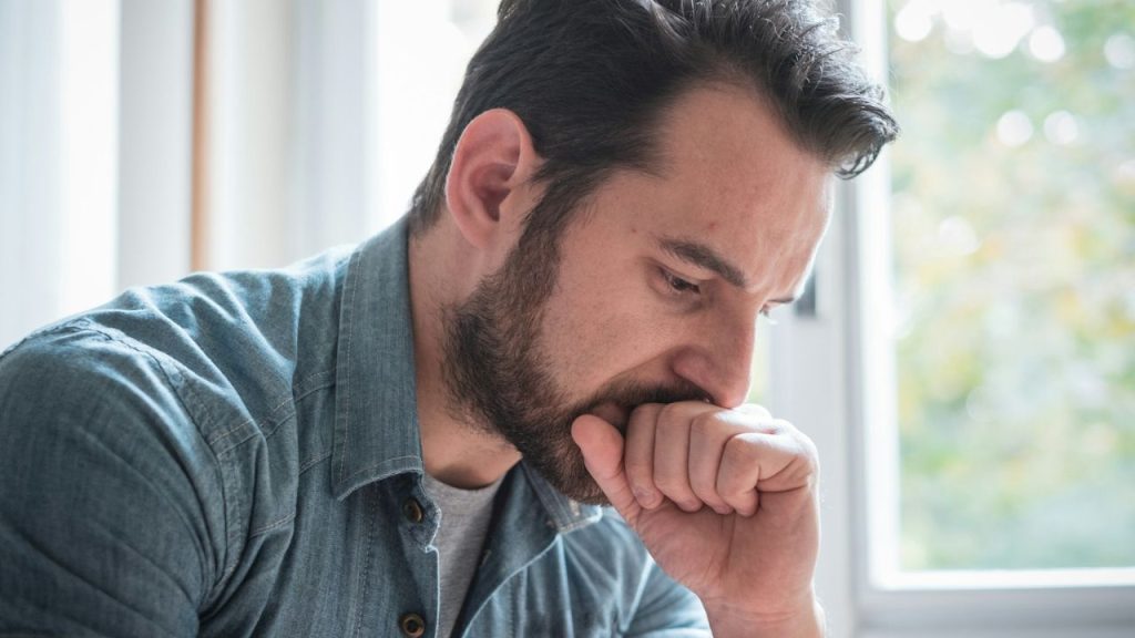 Close-up of a bearded man looking down and pensive, with his fist near his mouth.