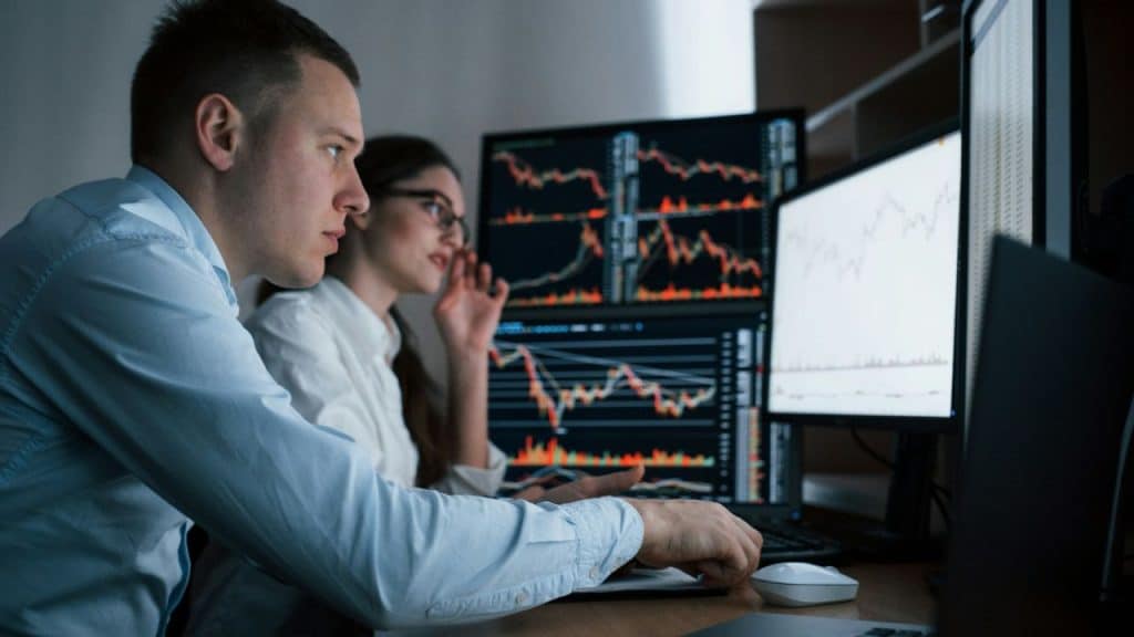 Two individuals intently watch multiple monitors displaying financial trading charts in a dimly lit office.