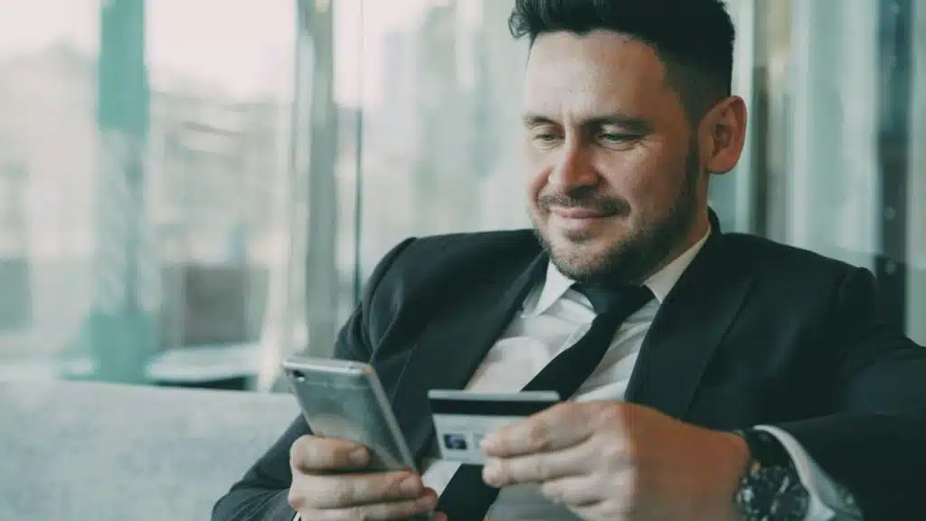 Smiling man in a suit holding a credit card and looking at a smartphone.