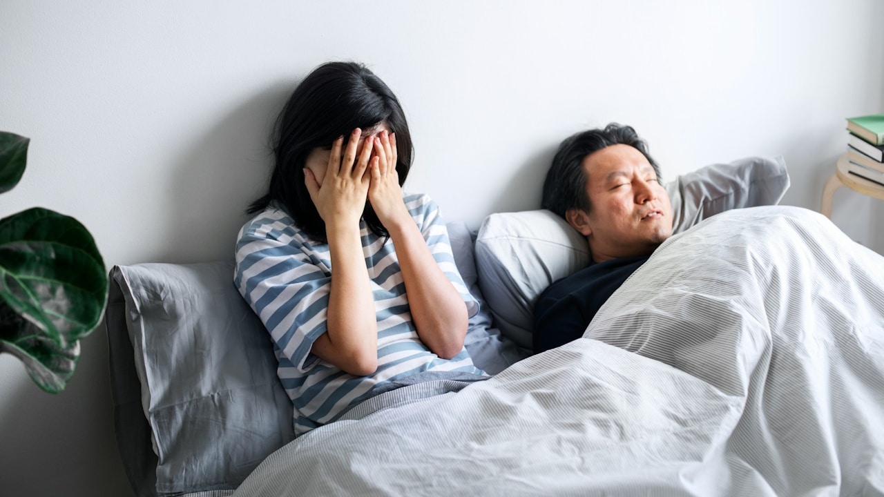 A distressed woman covering her face with her hands while a man sleeps next to her in bed.
