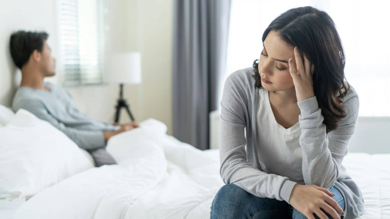 A woman with a furrowed brow sits on a bed, while a man faces away in the background.