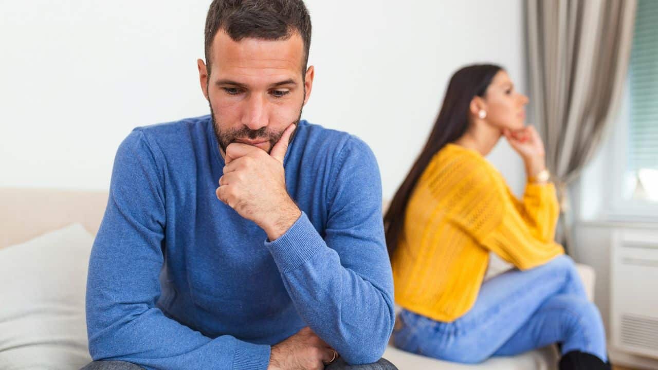 A man in a blue sweater sits contemplating, with a woman in yellow and blue blurred in the background.
