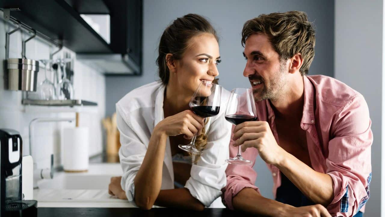 Couple in a kitchen looking at each other while raising glasses of red wine.