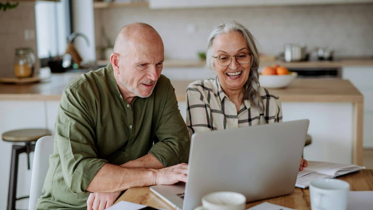 Happy older couple sitting at a kitchen table, looking at a laptop together.