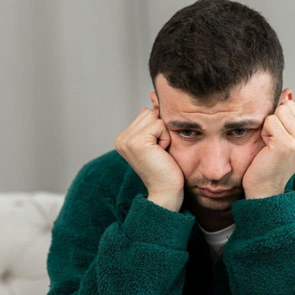 A sad-looking man in a dark green fluffy sweater resting his head in his hands.