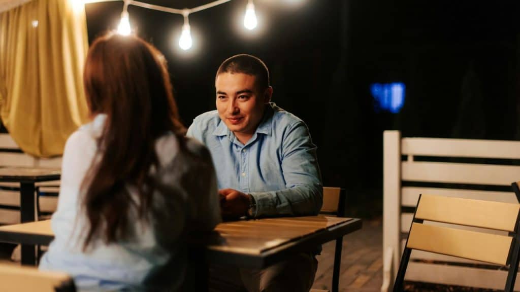A man sits at an outdoor table at night, smiling and listening to a woman opposite him.