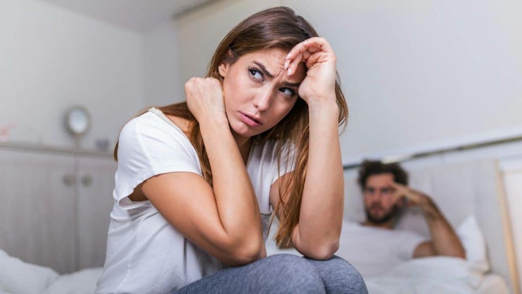 A distressed woman sits on a bed, holding her head, with a blurry man behind her.