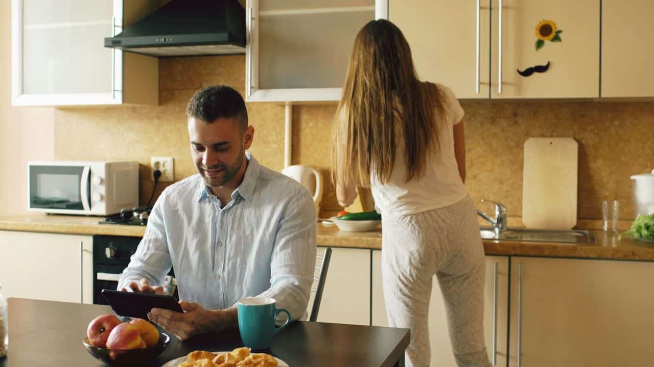 A man uses a tablet at a table while a woman works with food at a kitchen counter.