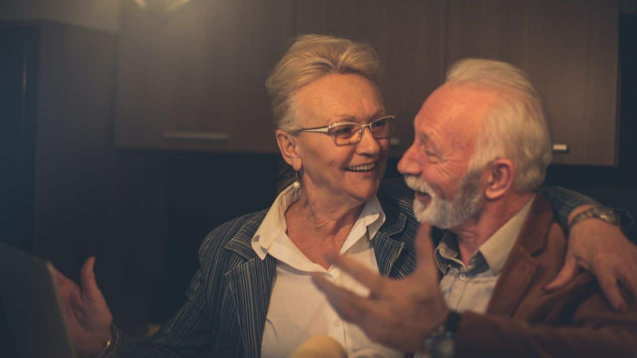 Smiling older woman in a blazer and turtleneck holding coffee cups opposite a man.