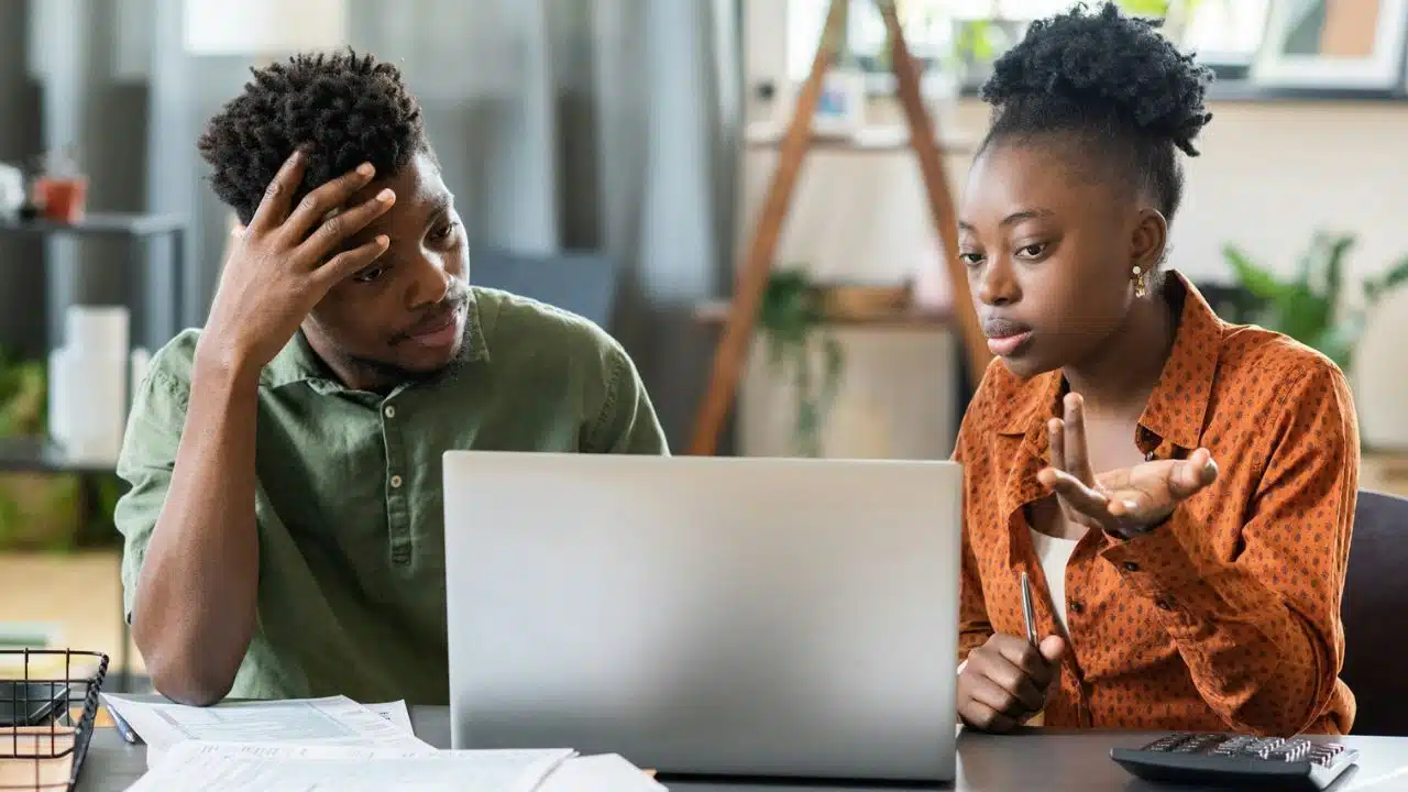 A Black man rests his hand on his head in frustration while a Black woman explains something, gesturing toward a laptop.