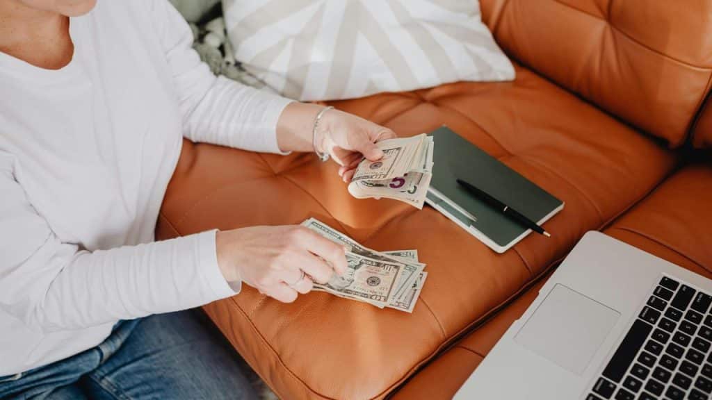 A person counting cash on a couch beside a laptop and notebook.
