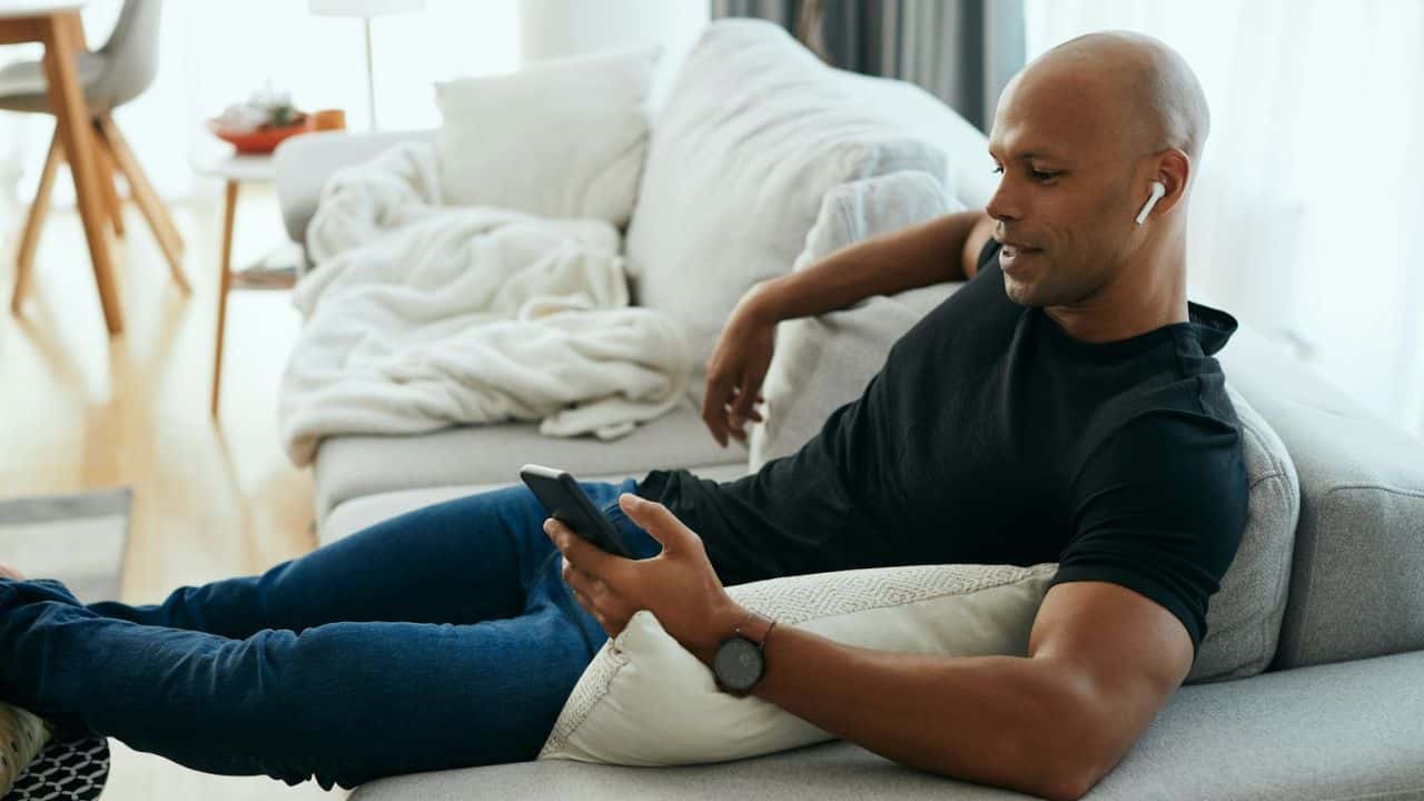 Bald man with earphones relaxing on a sofa, looking at his smartphone.