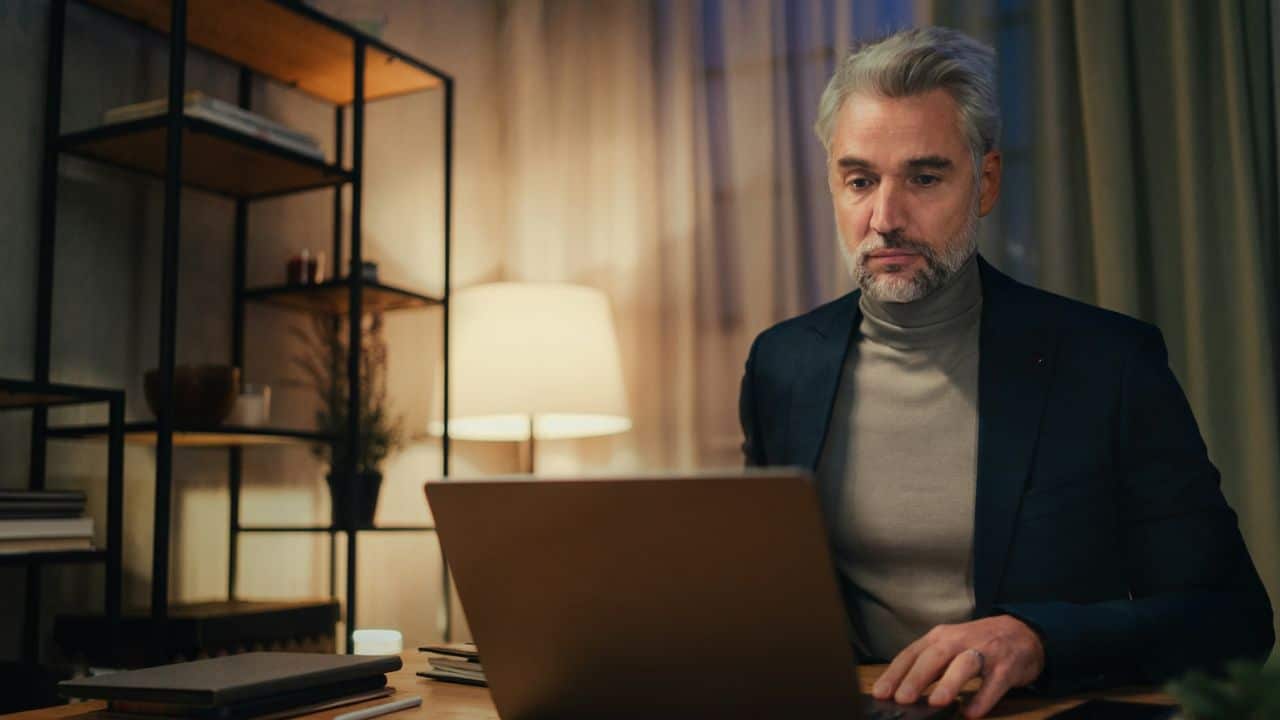 A mature woman looks down at a desk with a laptop and documents.
