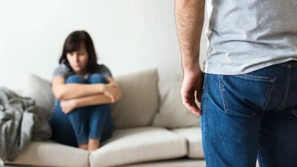 A woman is sitting on a couch with her hands folded across her legs as a man stands in front of her.