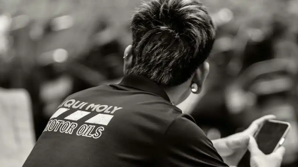 A person with short, spiked hair is seen from behind, wearing a shirt with the text “Liqui Moly Motor Oils.” They are holding a smartphone in one hand.