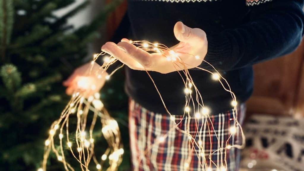 A person in a sweater and plaid pants holding strands of glowing fairy lights.