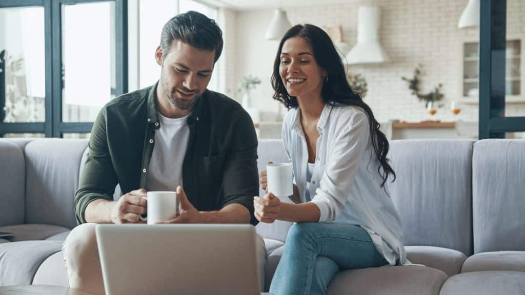 Couple looking down at a laptop while holding coffee mugs on a gray couch.