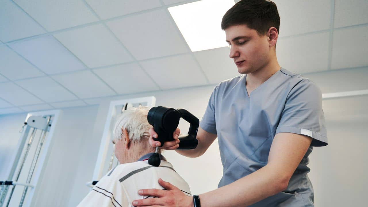A male practitioner using a black therapy device on an older person's shoulder.