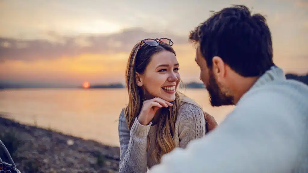 A cute smiling caucasian brunette sitting on the coast near a river with her loving boyfriend and flirting, in the background is sunset.