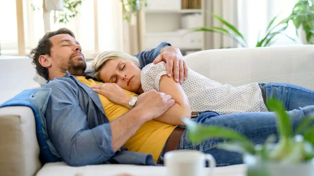 A man and woman cuddling on a white couch, both resting with their eyes closed.