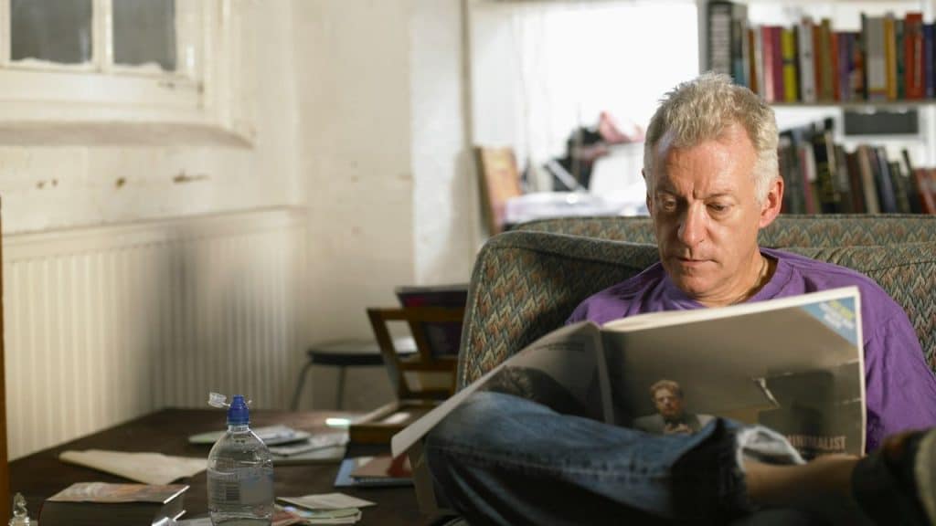 A middle-aged man sits and reads a magazine on a patterned armchair.