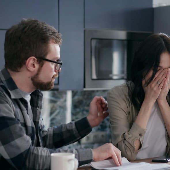 A distressed woman covering her face while a man comforts her and points to documents on a table.