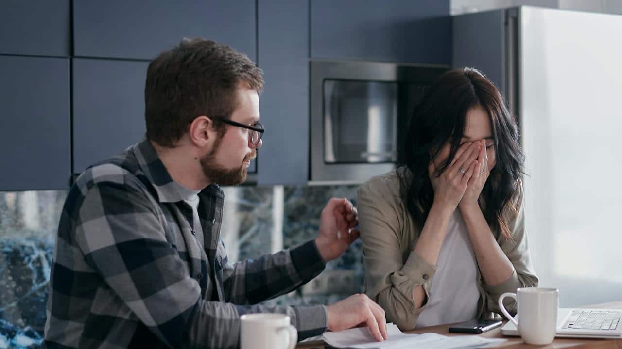 A distressed woman covering her face while a man comforts her and points to documents on a table.