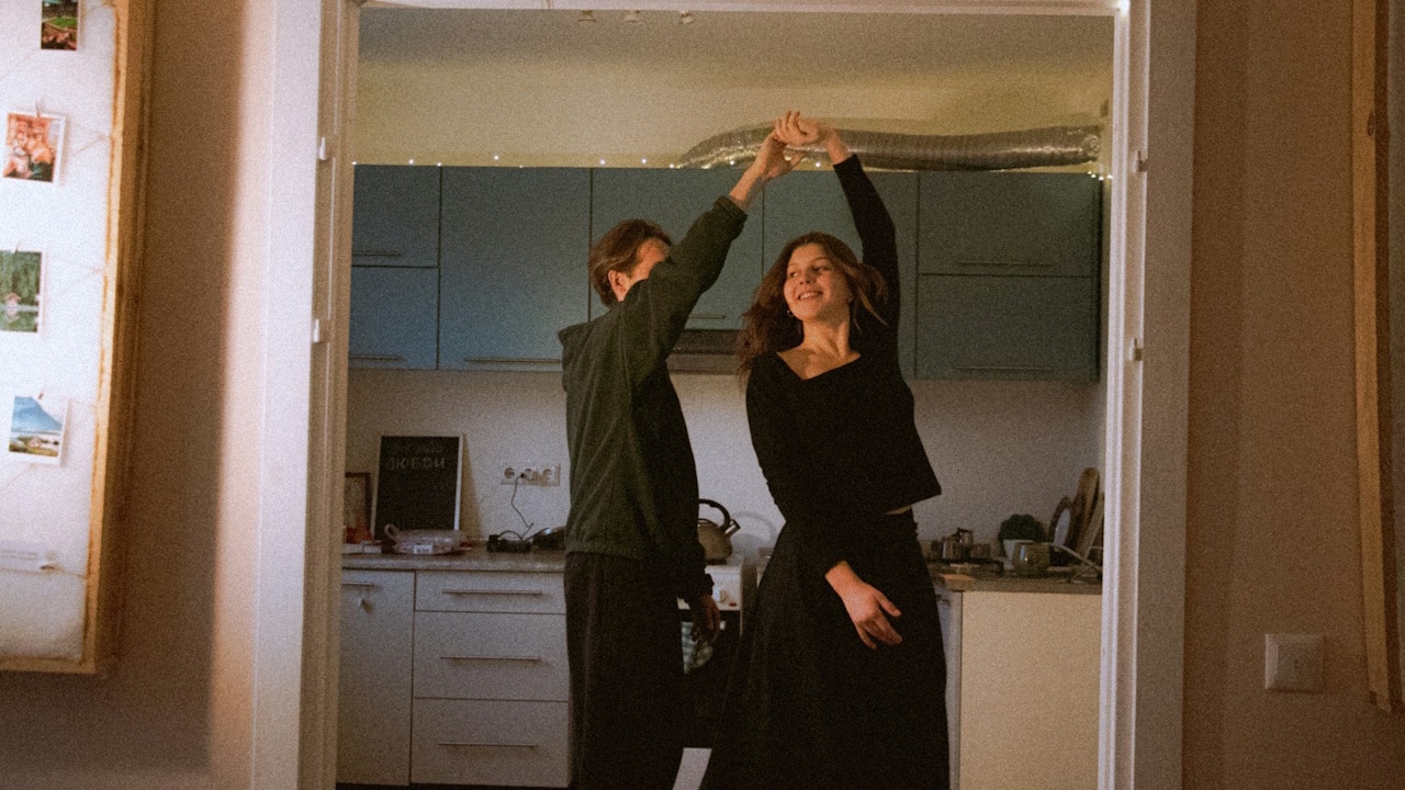 A happy couple is dancing in a doorway, holding hands over their heads, framed by holiday decorations.