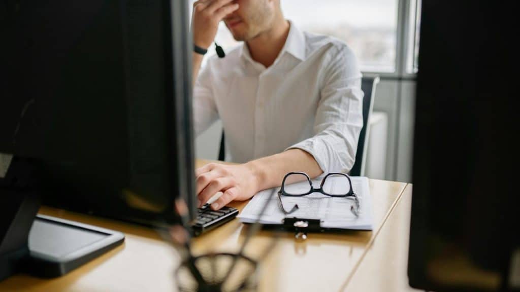 A man at a desk holding his face in frustration while working on a computer.