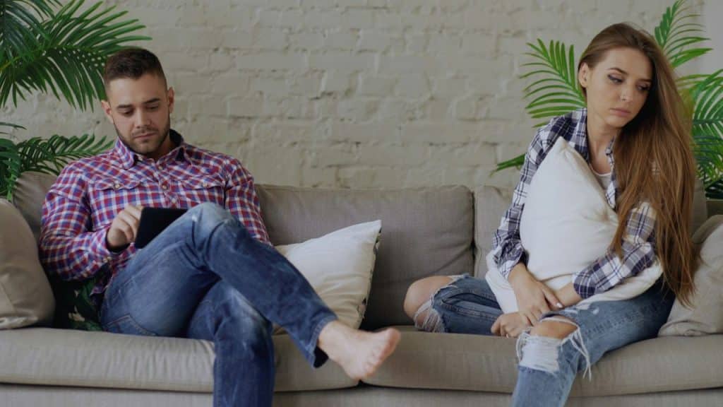 Man on a couch using a tablet; woman next to him holding a cushion.